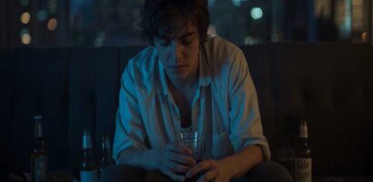 Jeune homme dans une pièce sombre, entouré de canettes de bière vides, tenant un verre, illustrant les dangers du binge drinking.